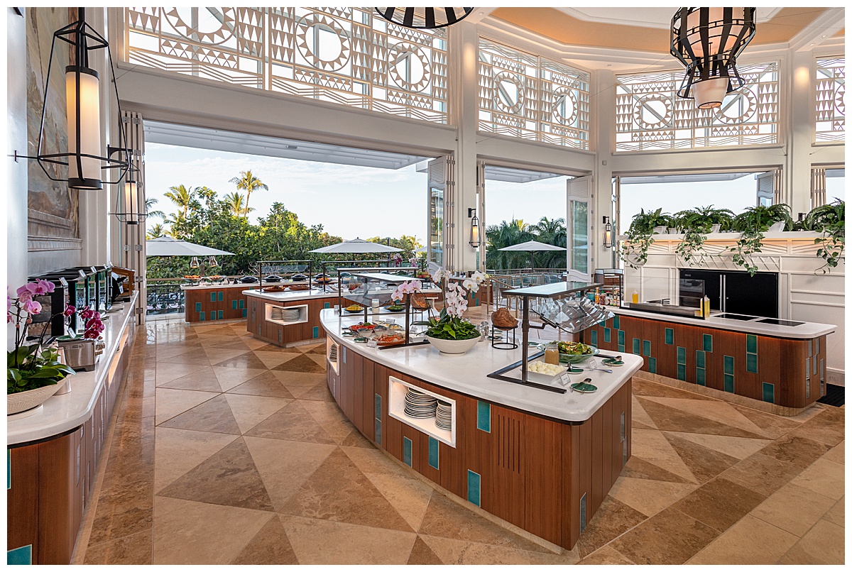 The buffet area of the Grand Wailea 'Ikena Restaurant. The white walls are scarce as the restaurant is very open air. The buffet tables are warm brown wood with white countertops. The floor is a brown marble triangle tile.
