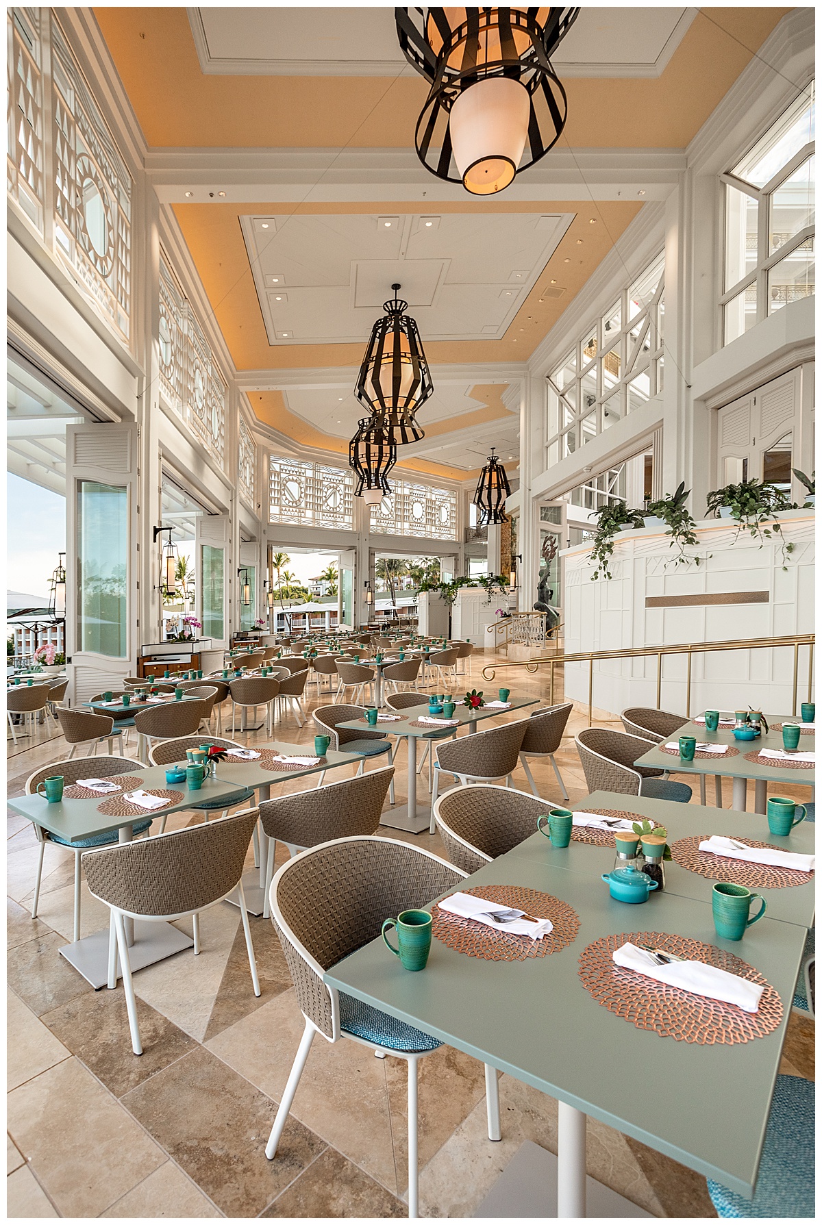The seating area of the restaurant. The white walls are scarce as the restaurant is very open air. The tables are light teal squares and rectangles, and the chairs are rounded with brown wicker backs. The floor is a brown marble triangle tile.