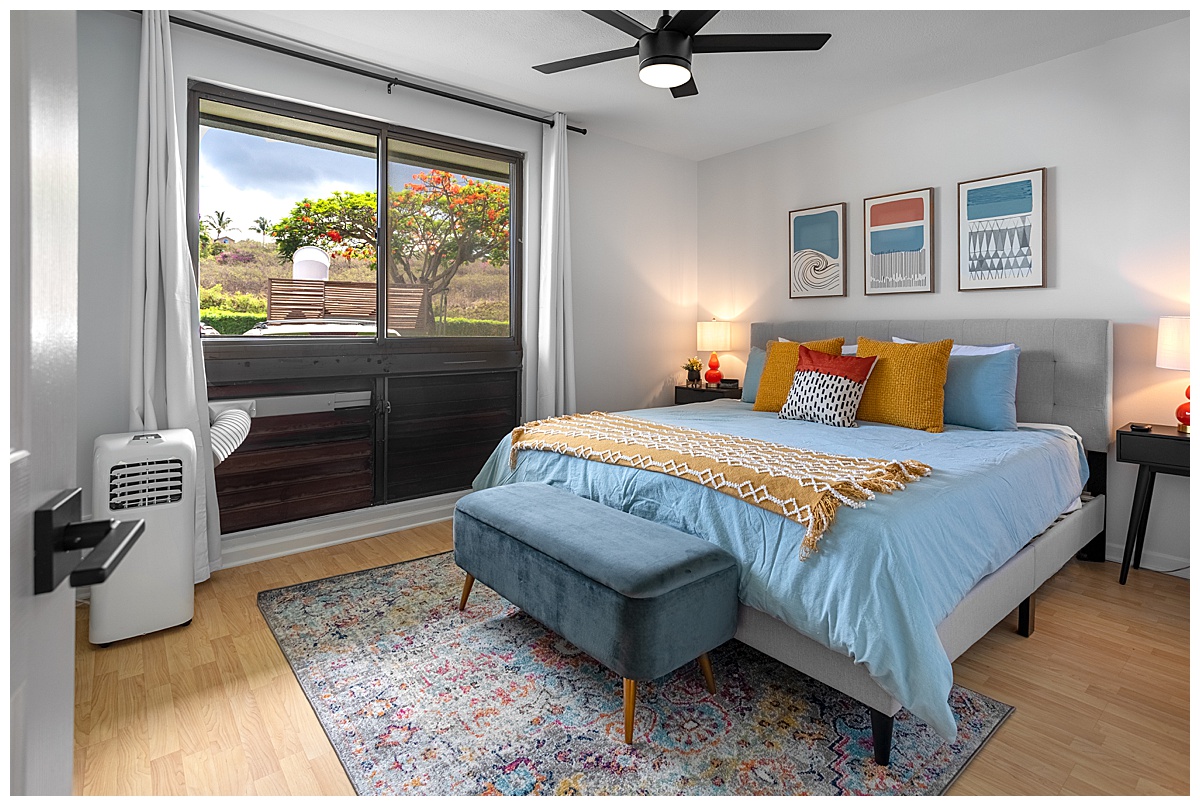 The bedroom of the vacation rental. The bedspread is blue with yellow, orange, and blue pillows. The walls are white and there are geometric paintings on the wall. There is a ceiling fan and window.