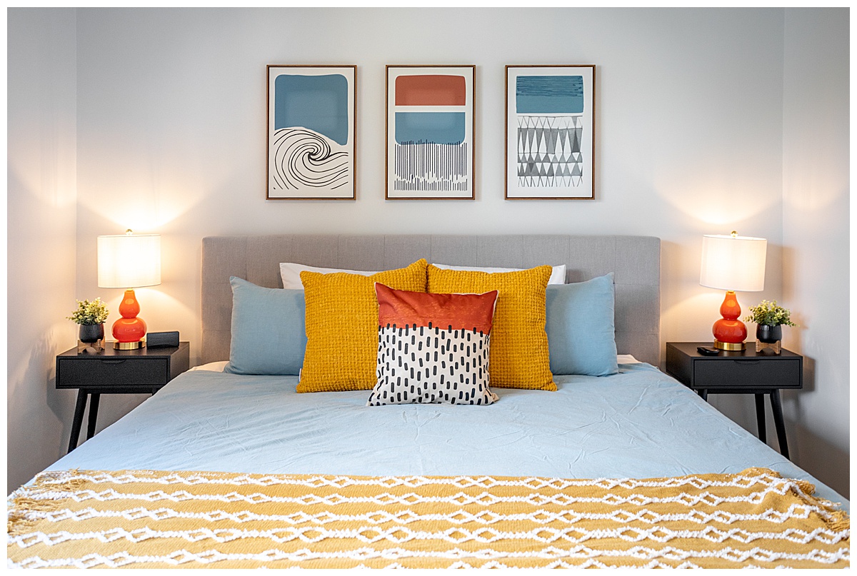 The bedroom of the vacation rental. The bedspread is blue with yellow, orange, and blue pillows. The walls are white and there are geometric paintings on the wall.