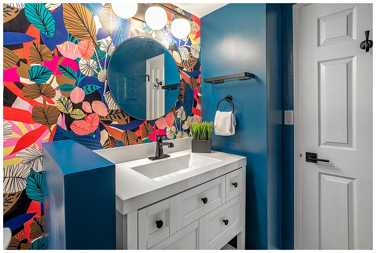 The bathroom has a bright colorful pattern on one wall. The pattern is leaves of all different colors, and the other walls are bright teal blue. The sink is white and there is a round mirror above it.