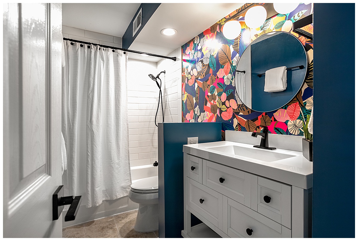 The bathroom has a bright colorful pattern on one wall. The pattern is leaves of all different colors, and the other walls are bright teal blue. The sink is white and there is a round mirror above it. The shower is all white in the background.