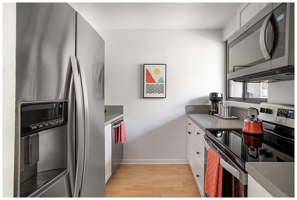 The kitchen is simple with white cabinets, gray countertops, white walls, and stainless steel appliances. There are small orange decor pieces.