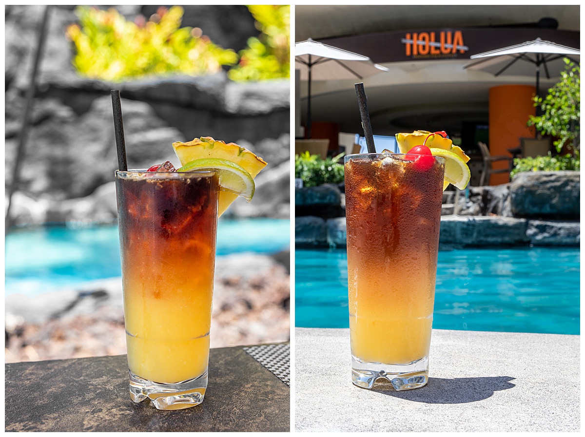 A mai tai on the side of the pool from the poolside bar. The drink is in a tall glass with yellow on the bottom and dark rum on top. It is garnished with a pineapple, lime, and cherry. The pool and poolside bar, Holua, are in the background.