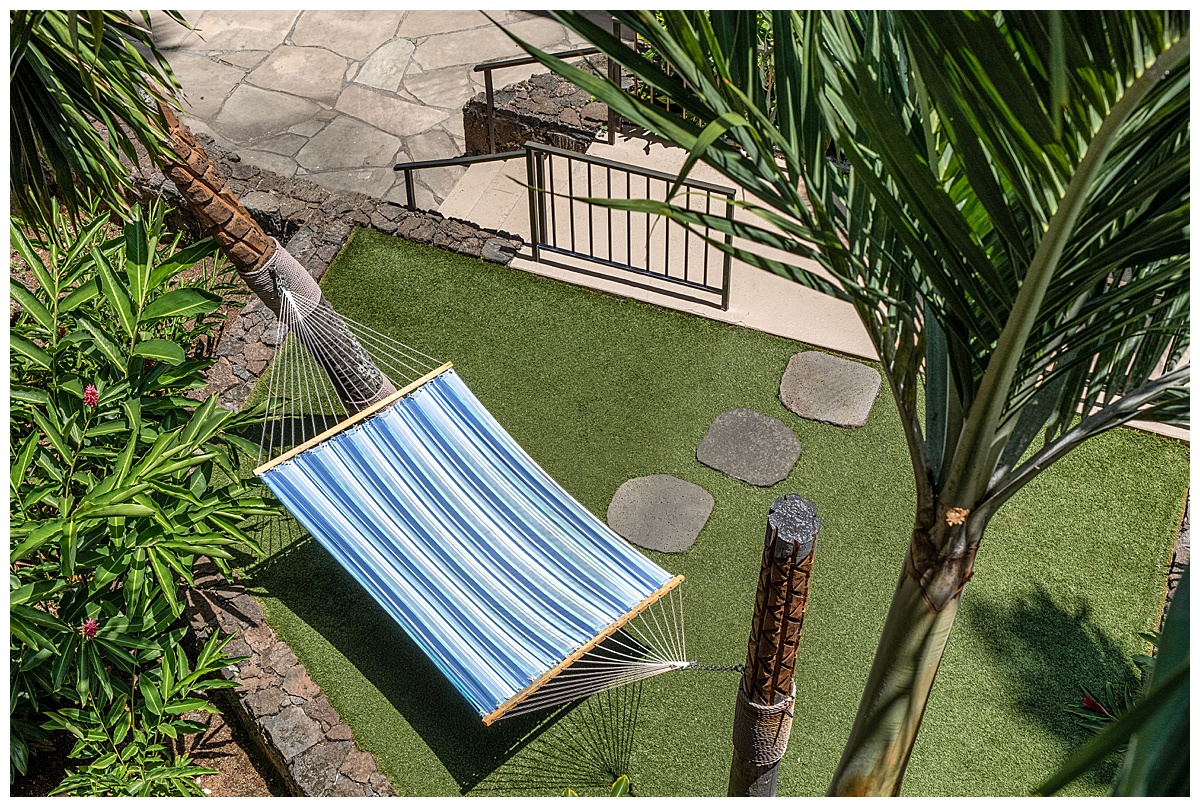 A closeup of the hammock swing next to the adults only pool. The hammock is blue and white stripes and it's over green grass surrounded by foliage.