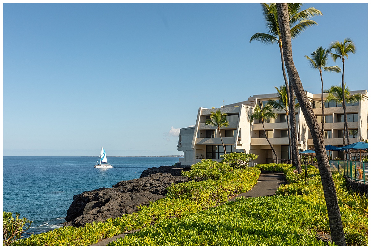 A view of the Outrigger Kona Resort & Spa from the oceanfront path. A sailboat is sailing past on the water.
