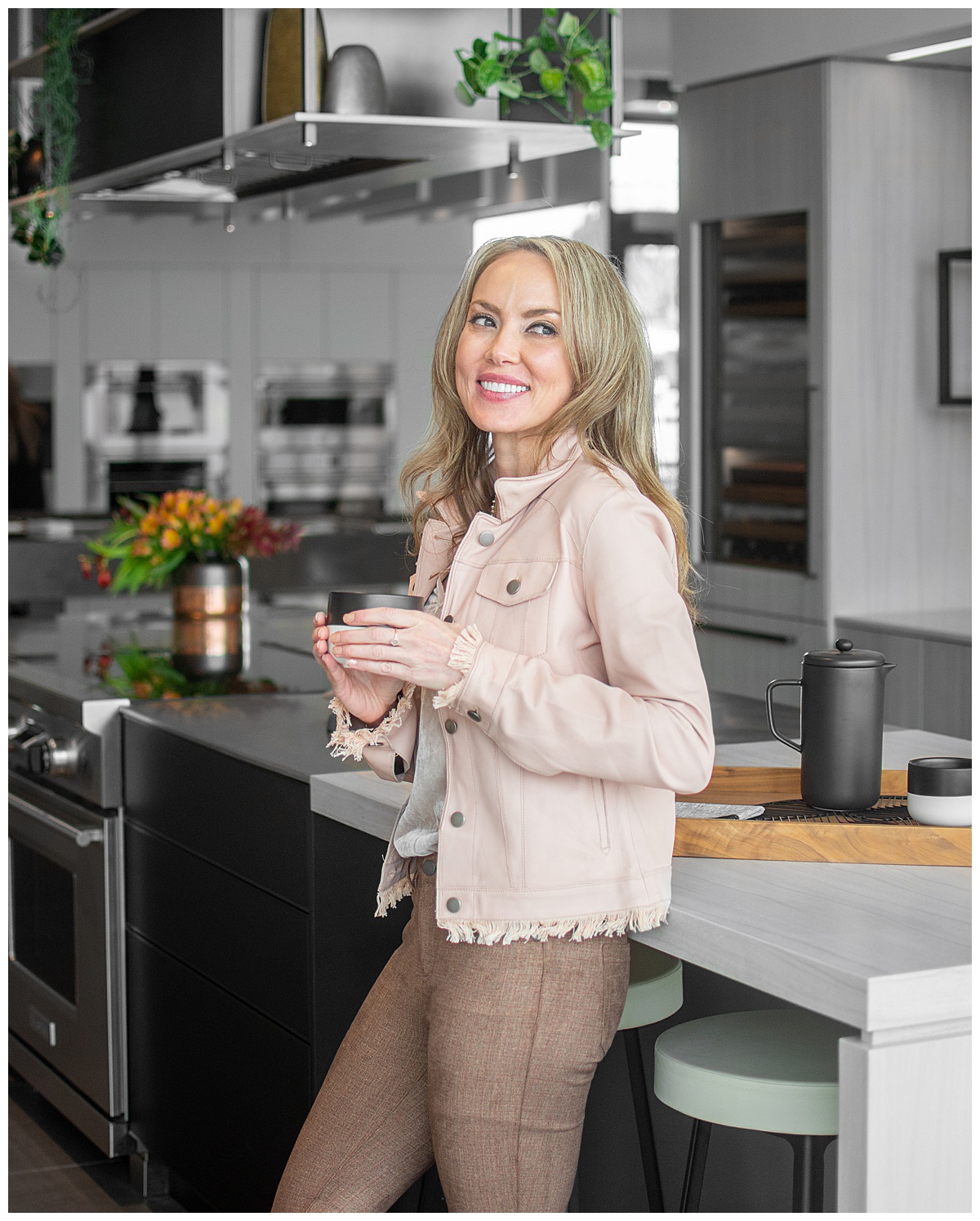 A woman stands in a kitchen showcasing her interior design. She is wearing a pink jean jacket and drinking from a cup. The kitchen is gray with stainless steel appliances and greenery accents.