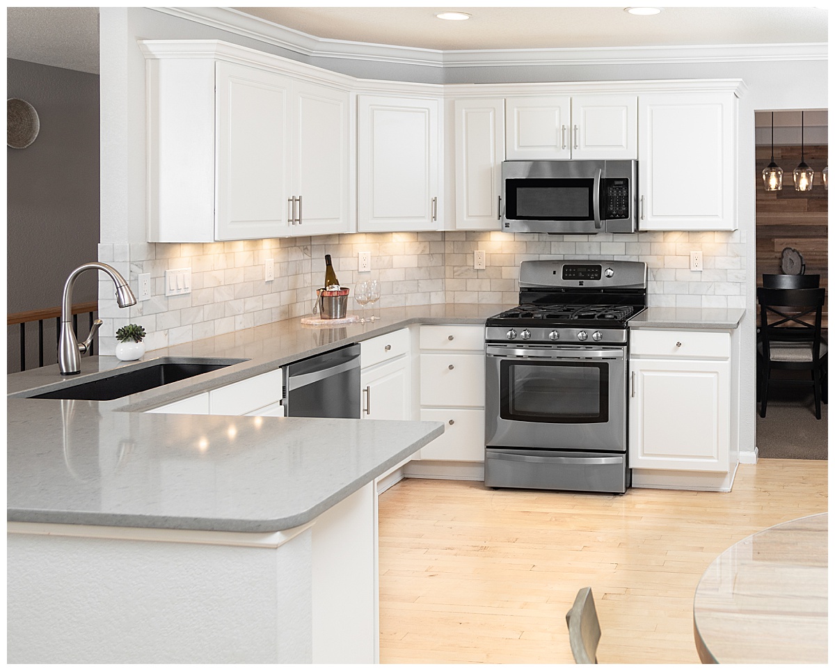 A full photo of a kitchen showing the sink, oven, and general layout. The kitchen has white cabinets and a white/gray stone tile backsplash. The floor is a light wood.