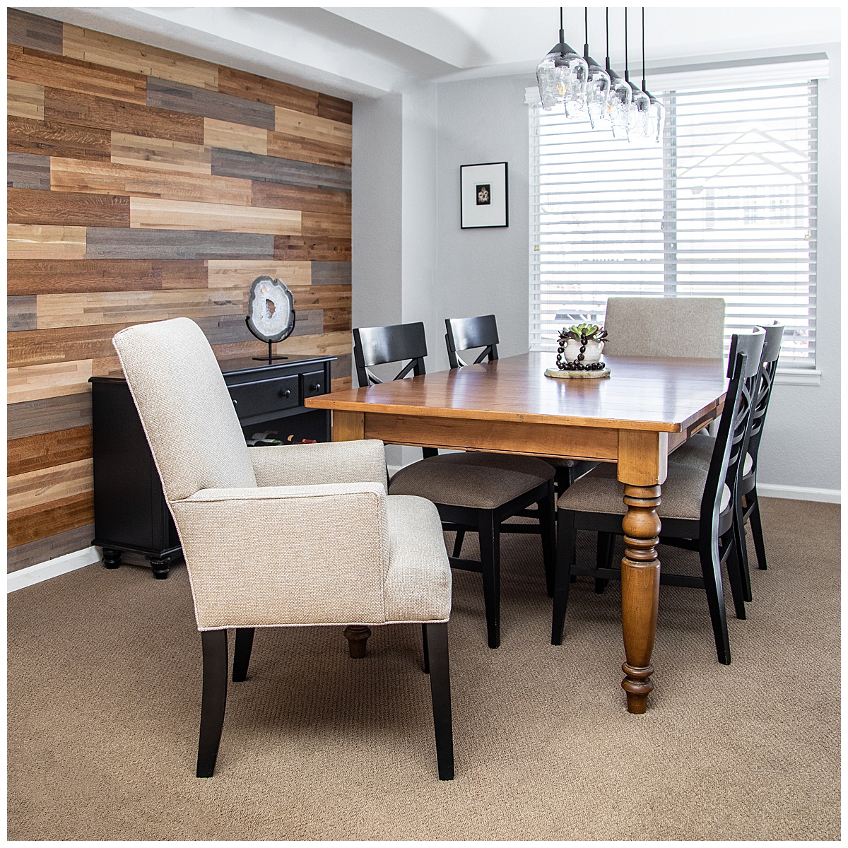 A dining room with gray walls and a wood accent wall. The floor is beige carpet and there is a window on the back wall. The table is brown finished wood, the chairs are black wood, and there is a head chair that is plush and cream colored.